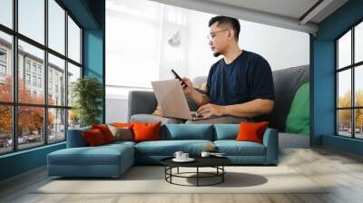 Asian man sitting on couch at home using laptop and cell phone Wall mural