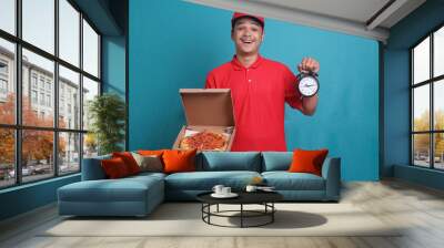 Delivery worker in red uniform is holding boxes of pizza and a clock. On time fast food delivery concept. Wall mural