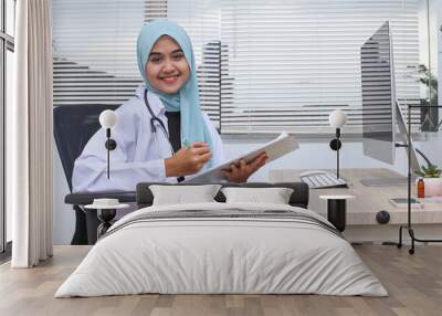 Muslim woman doctor writing on clipboard while using laptop on desk in hospital. healthcare and medical occupation concept. Wall mural