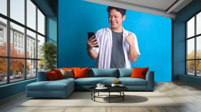 Smiling happy Asian man using smartphone while clenching fist celebrating victory Wall mural