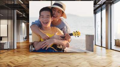 Asian special child on wheelchair is singing, playing ukulele happily on the beach with parent,Natural sea beach background,Life in the education age of disabled children,Happy disability kid concept. Wall mural