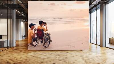 Asian special child on wheelchair on the beach with parents in family holiday to travel, Take photo the camera nature around the sea beach, Life in the education age, Happy disabled kid concept. Wall mural