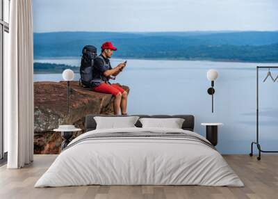 hiker man sitting and using smartphone on edge of cliff, on a top of the rock mountain Wall mural