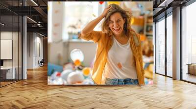 A joyful woman celebrates amidst a chaotic and colorful party scene with balloons, confetti, and decorations, conveying happiness and excitement in a messy environment. Wall mural