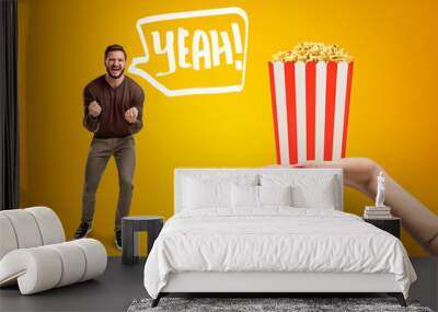 Young man in casual clothes shouting 'Yeah' and holding clenched fists because he is so happy about pop corn box he's neen offered on amber background. Wall mural