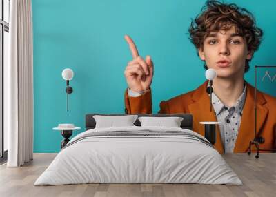 Young man in smart casual attire pointing upwards on solid blue background, with a curious look Wall mural