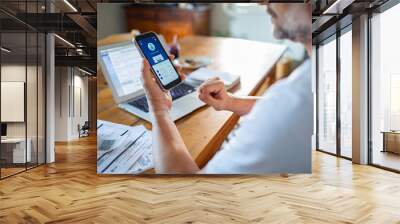 Man checking online bank account balance on smartphone while working on laptop at home Wall mural