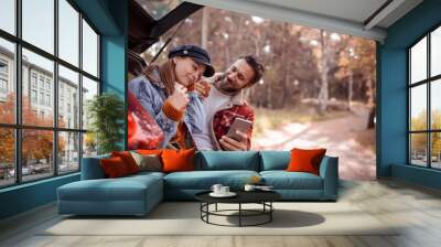 Young couple eating sandwiches and using a smartphone while sitting on a car trunk in a park Wall mural