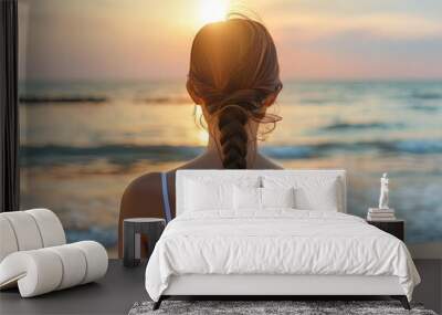 Woman with a braid enjoying a peaceful sunset by the ocean, reflecting and relaxing on the beach, with the sun glowing on the horizon. Wall mural