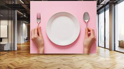 Female hands holding a fork and a spoon next to an empty plate on a pink background closeup top view : Generative AI Wall mural