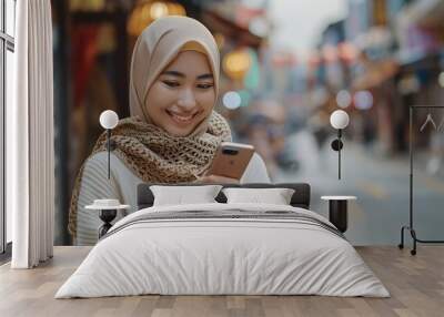 Portrait of smiling young asian muslim woman in cream sweater and hijab using mobile phone pointing finger at screen phone blank screen People islam religious concept : Generative AI Wall mural