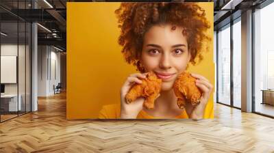 A young woman with curly hair smiling as she holds two fried chicken legs in front of her face against a yellow background. Wall mural