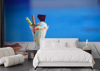 Panorama of glass with rainbow ice cream and colorful syrup on blur bokeh background with copy space. Waffle chocolate cones on white table top of blue water pool Wall mural