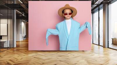 Portrait of a little girl in an oversized adult blue blazer, stylish red glasses and a round brown hat. Child wearing adults clothes. Isolated on pink background. Wall mural