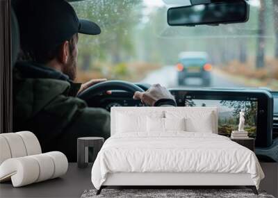 A man in his mid-30s is driving an SUV through a scenic mountainous landscape wearing casual attire and a cap Wall mural