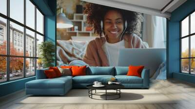 Beautiful young African American girl with headphones and laptop sitting with folded legs on the floor in a cozy room. Smiling black female student learning online. Remote education concept. Wall mural