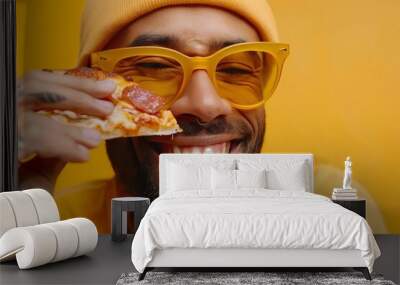 Close-up portrait of a funny bearded Caucasian man in yellow glasses, T-shirt and knitted hat eating pizza slice on yellow background. Cheerful guy laughing and squeezing his eyes. Wall mural