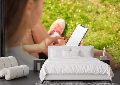 Cute teen girl looks at the screen of her smartphone sitting on a green lawn in the garden. Close-up phone display. Wall mural