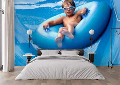 Joyful boy in swimming goggles slides down the blue water slide on an inflatable boat in the water park, water splashes, joyful mood, summer vacation Wall mural