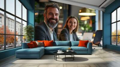 Two business professionals smiling in a modern office setting with laptops and warm lighting Wall mural