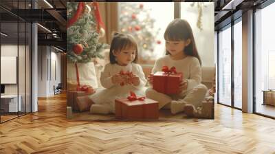 Two young girls sitting by the Christmas tree, exchanging gifts and embracing the joyous holiday spirit in a cozy living room setting Wall mural