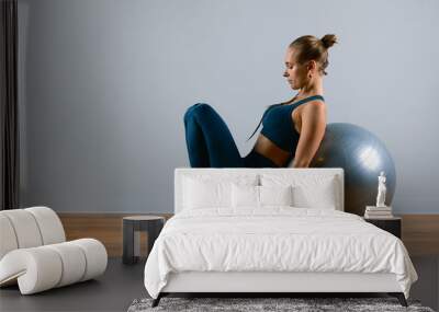 Young, beautiful, athletic girl doing exercises on fitball in the gym on a gray background. Sporting Slavic girl in a blue, green suit. Copy space, gray background, sport banner for advertising. Wall mural