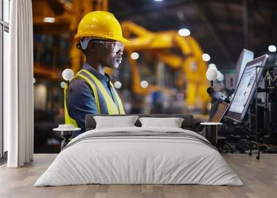 Young cheerful African American female engineer, technician or worker on a factory floor. Confident black woman in safety helmet and vest operates complex industrial equipment, setting up the machine. Wall mural