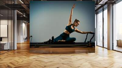 Young girl doing pilates exercises with a reformer bed. Beautiful slim fitness trainer on reformer gray background, low key, art light. Fitness concept Wall mural