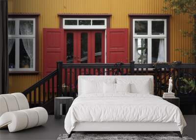 Picturesque facade of a traditional wooden house in yellow on the wooden exterior, burgundy windows, and red door. Two bicycles are resting on the balcony.  Reykjavik, Iceland  Wall mural