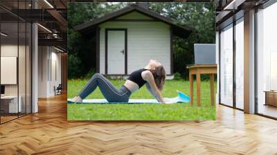 Teenage girl does exercises watching online tutorial on a laptop outdoors Wall mural