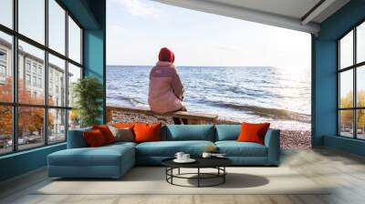 girl sitting on a bench against the background of an evening sunset looks at the water Wall mural