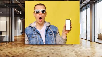 Stylish Caucasian man in a jeans on a yellow background with 8 bit glasses. Copy space. Holds a smartphone with a shock on his face. Wall mural