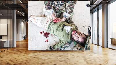 Soldiers trying to stop bleeding at wounded comrade who lying on floor, suffering and screaming in paine. Commando fighter pressing with hands on bloody wound at friends stomach, giving emergency care Wall mural