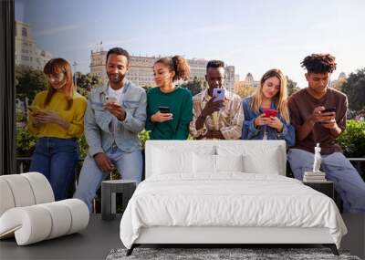 Group of multicultural friends sitting outside using mobile phone. Serious people addicted to technology, sitting outdoors in a row with cellphones in hands. Wall mural