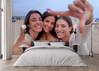 Happy three female friends on beach travel together, looking at camera smiling and gesturing a frame with fingers. Caucasian women enjoying summer in california. Wall mural