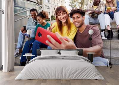 Multiracial young friends sitting on stairs hanging out, using phone to take photos and send messages while being outside enjoying the day. Millennial generation. Wall mural