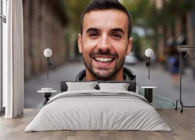 Portrait of a young handsome man smiling and looking at camera. Happy male wearing headphones in street on the city. Close up of a cheerful millennial caucasian guy outdoors. Wall mural