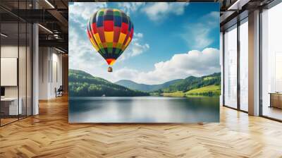 Colorful hot air balloon flying over a lake. Beautiful travel landscape with green hills, blue sky, & white clouds on a sunny day. Copy space.  Wall mural