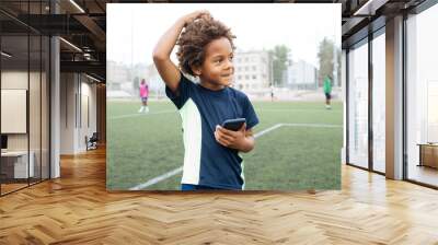 Smiling and excited black boy showing smartphone empty screen. Looking to camera. Cellphone display mockup mobile app advertisement. African american kid in football field. Blank screen Wall mural