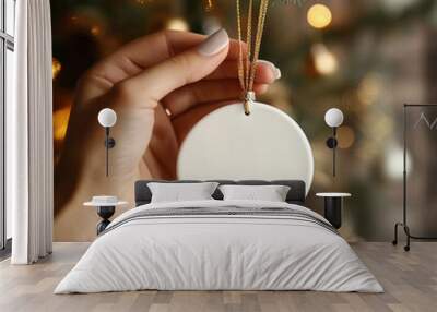 A white round Christmas ornament held by a hand against a bokeh background of a Christmas tree. Wall mural