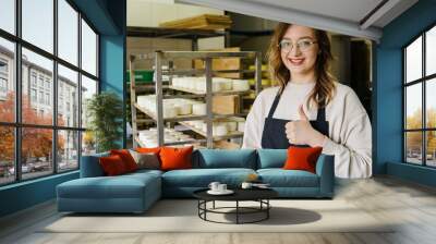 Young smiling woman standing at shelf with cheese heads and showing thumb up. Wall mural