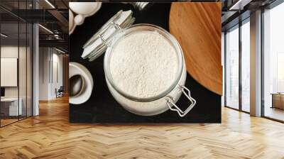 Open glass jar with white wheat flour on black wooden table among other kitchen stuff Wall mural