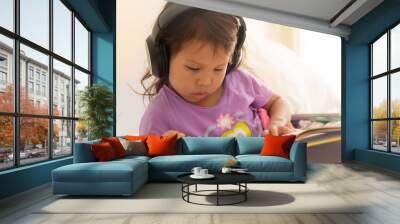 A little girl reading a book at home and listening to music wearing headphones. Audio books. Wall mural