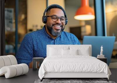 A cheerful man wearing glasses and headphones enjoys a fresh salad while working on his laptop in a modern, well-lit office space. His broad smile suggests a pleasant and engaging work environment Wall mural