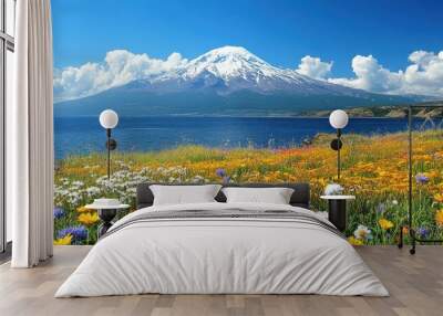 A snow-capped mountain peak rises above a field of wildflowers along a blue lake, with white puffy clouds in the clear sky. Wall mural