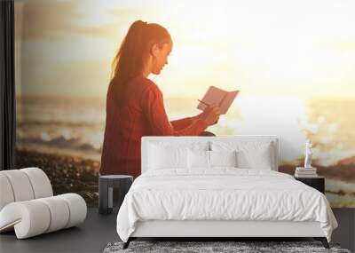 A young, happy woman listening to music and writes her thoughts in a personal diary on the beach at sunset. Enjoy your time and moments. Travel and relaxation Wall mural