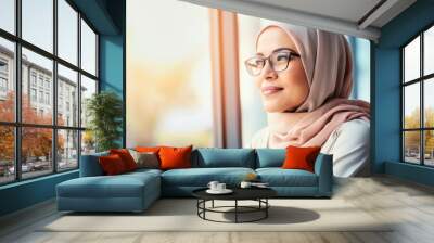 Beautiful satisfied cheerful rejoiced smiling young muslim woman wearing hijab and glasses sitting by the window, multinational cultures people Wall mural