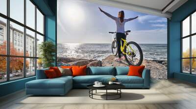 Free woman with bicycle stands on the stones on the seashore and raises hands to the sky as a sign of freedom or victory. Female with open hands. People with active and sporty lifestyle Wall mural