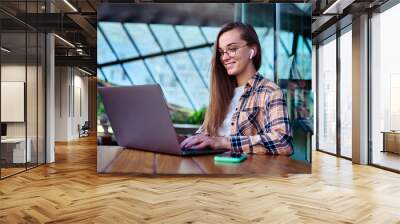 Happy woman freelancer in wireless headphones and eyeglasses using a laptop for remotely working online in cafe. Modern people with digital smart mobility lifestyle Wall mural