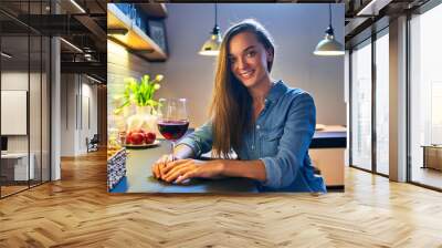 Portrait of beautiful cute young casual smiling happy drinking woman holding a glass of red wine and sitting at the table in the loft style kitchen at home Wall mural